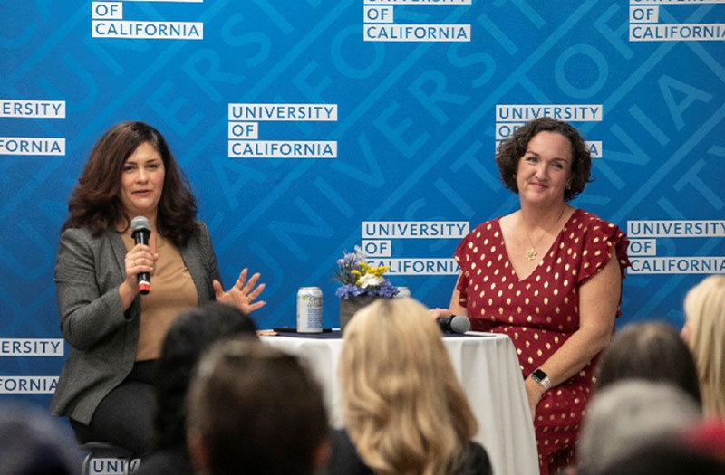 Two women seated on a stage at an event with a blue backdrop.