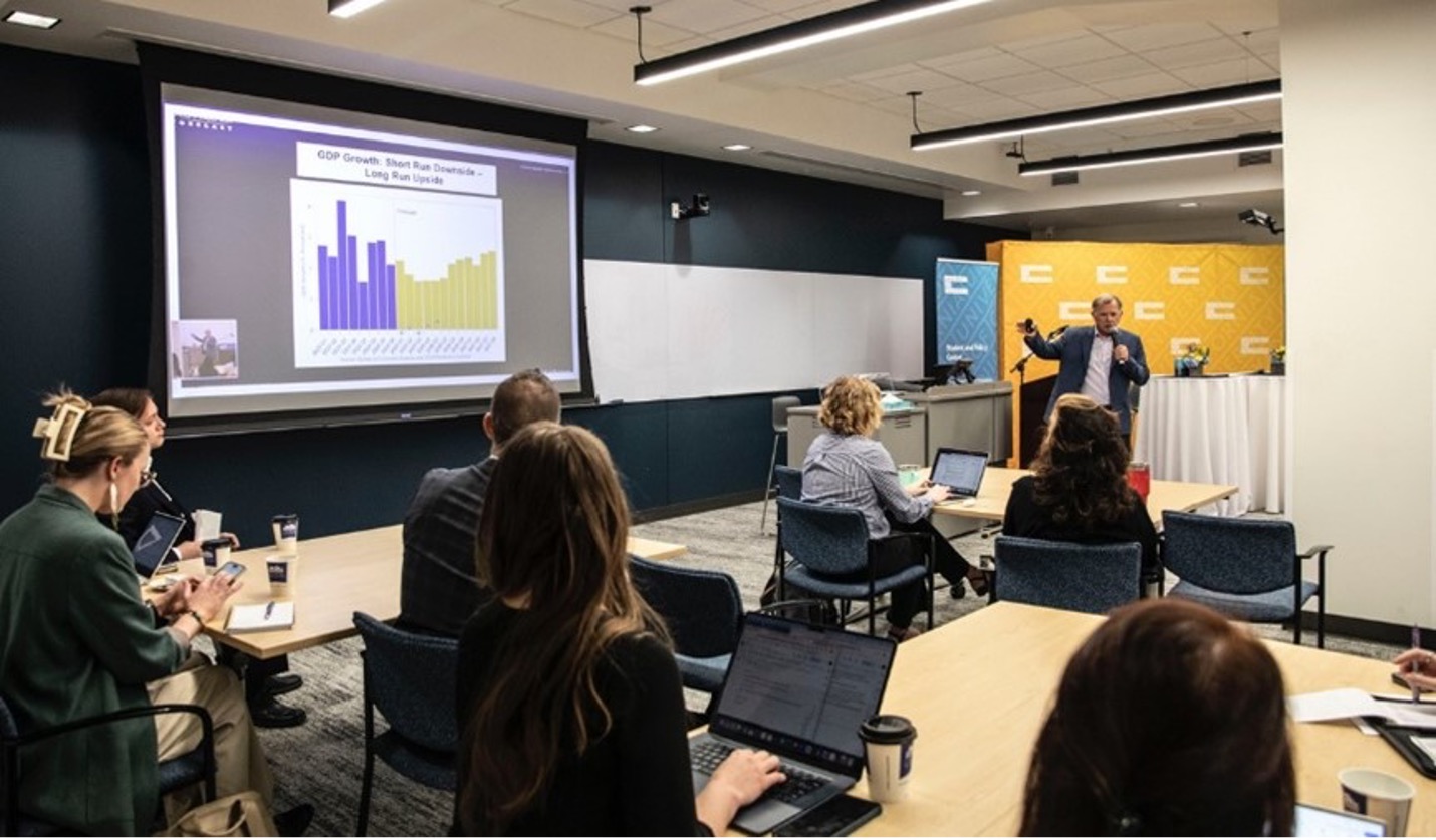 A presenter in a conference room points to a bar chart on a screen.