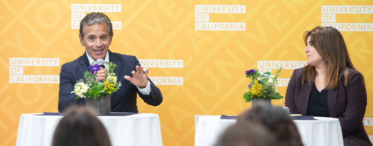 Two people seated at tables speaking with a University of California banner backdrop.