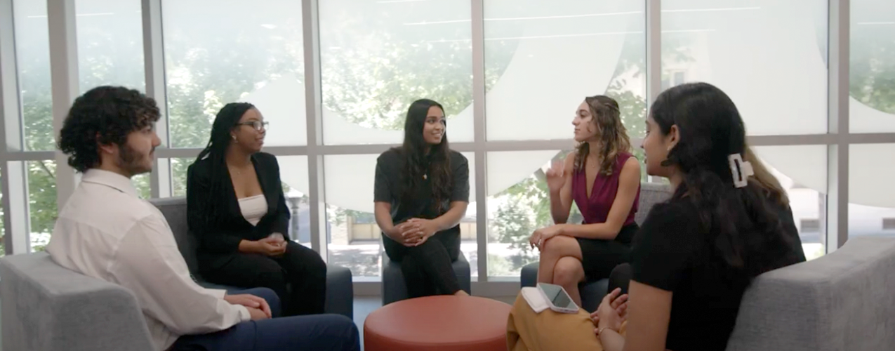 Group of five people sitting in a lounge area talking, with large windows in the background.